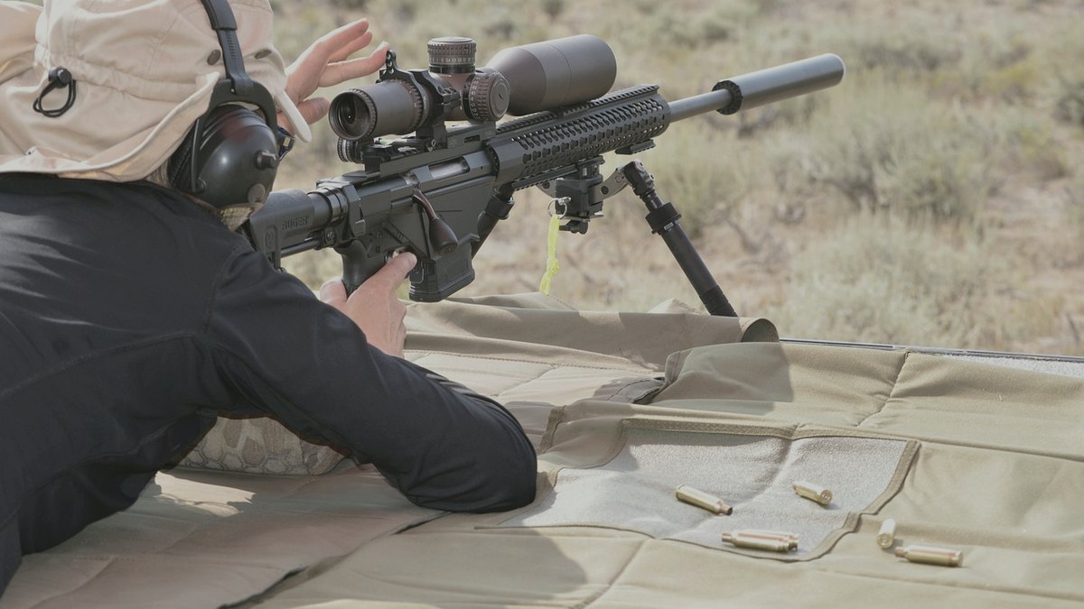 Shooter prone behind a scoped rifle fitted with a suppressor
